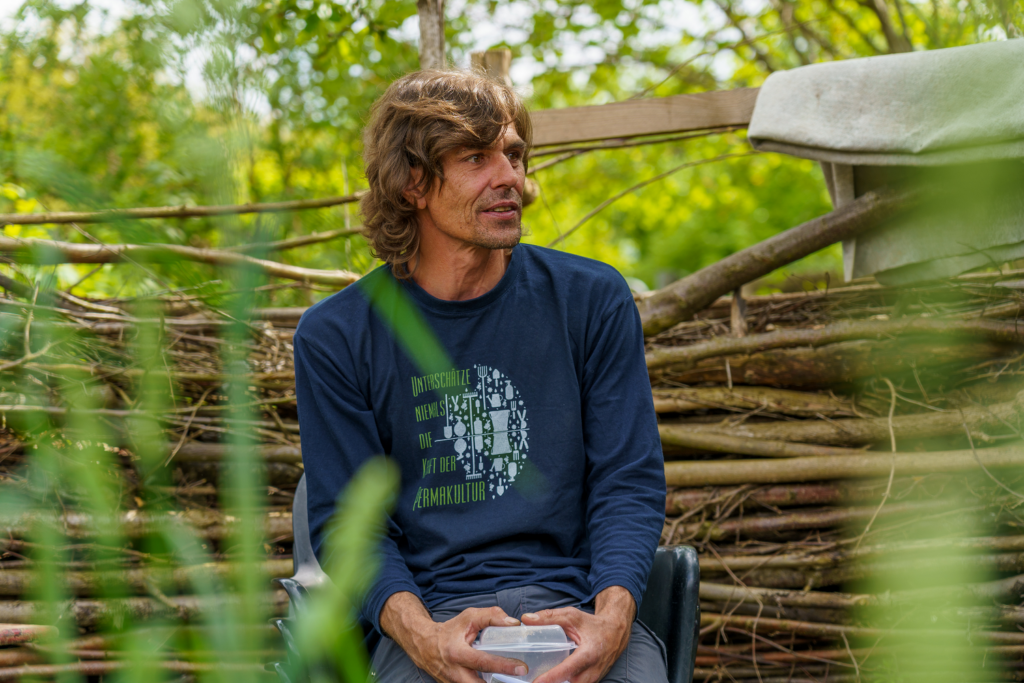 Emanuel Schmid sitzt in einem naturnahen Sofagarten mit Holz, Pflanzen und grünem Hintergrund.